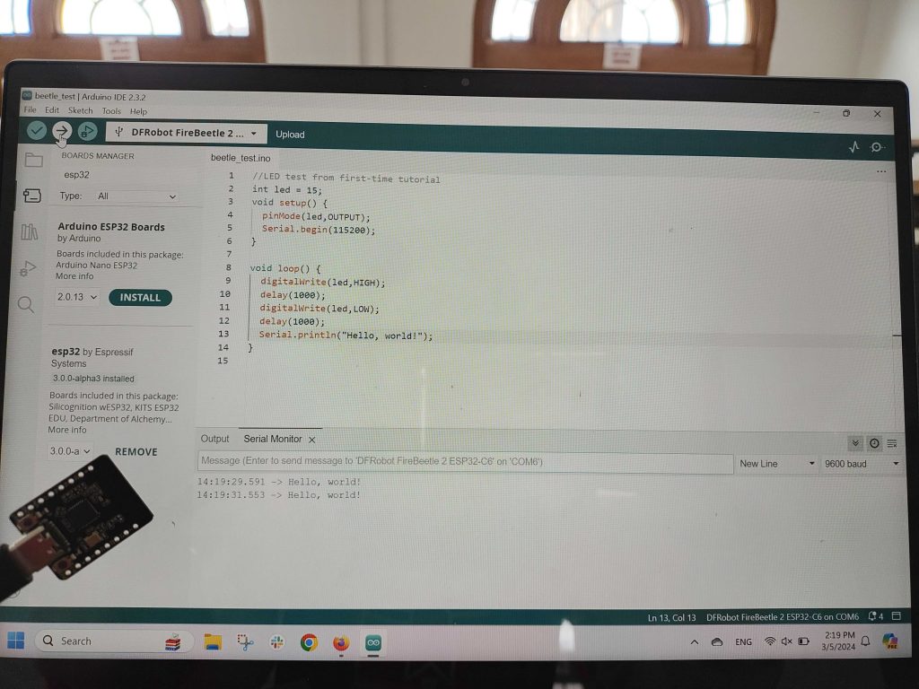 A Beetle with USB connection is held in front of a screen displaying the Arduino IDE, where the Serial Monitor reads "Hello, world!" twice.